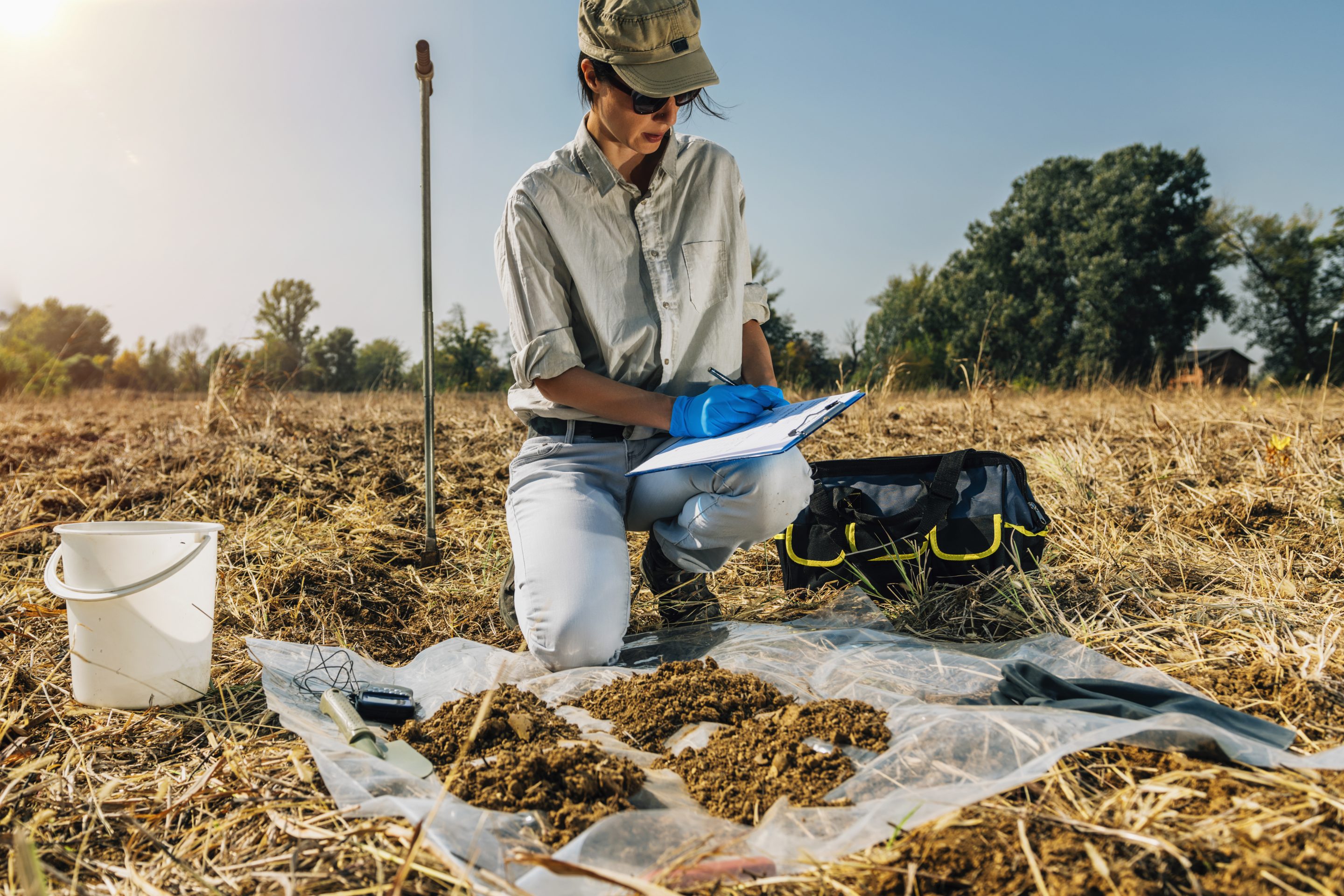A womain writing notes on a clipboard during a soil survey in an open field.